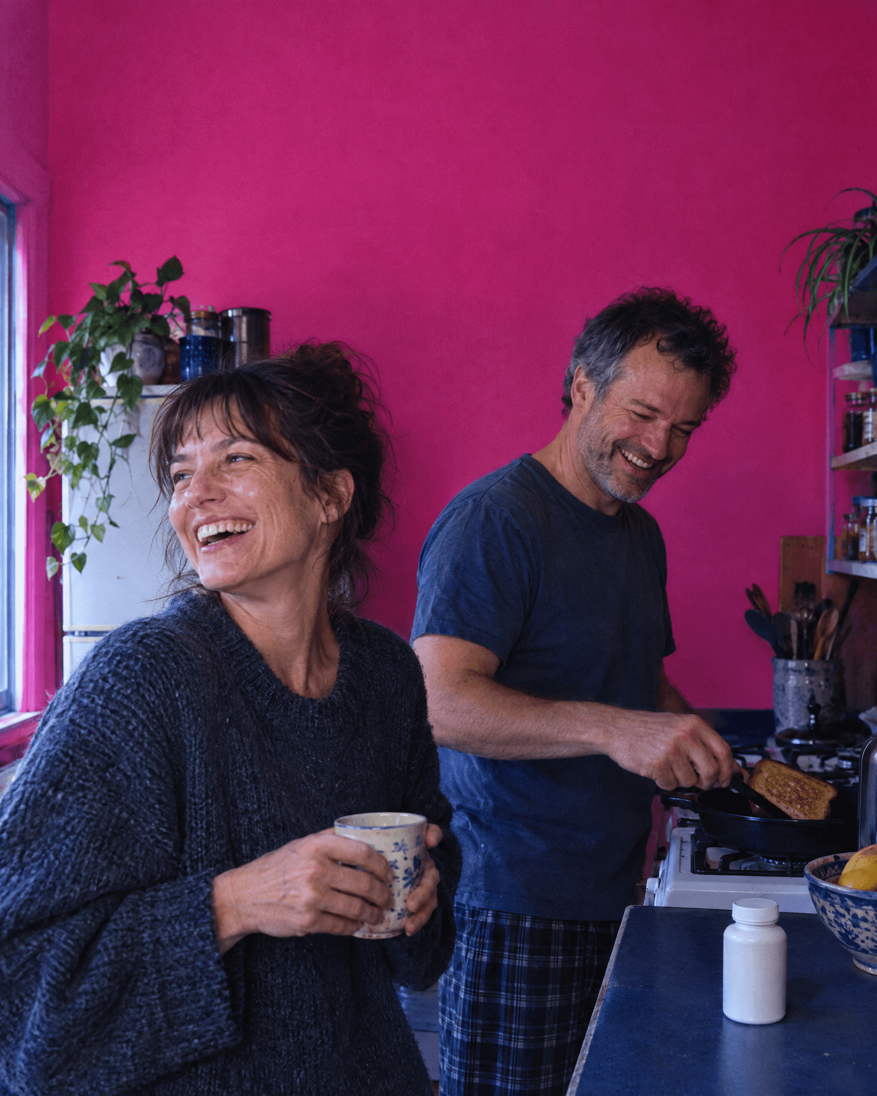 Two adults laughing in a pink-walled kitchen while making toast.