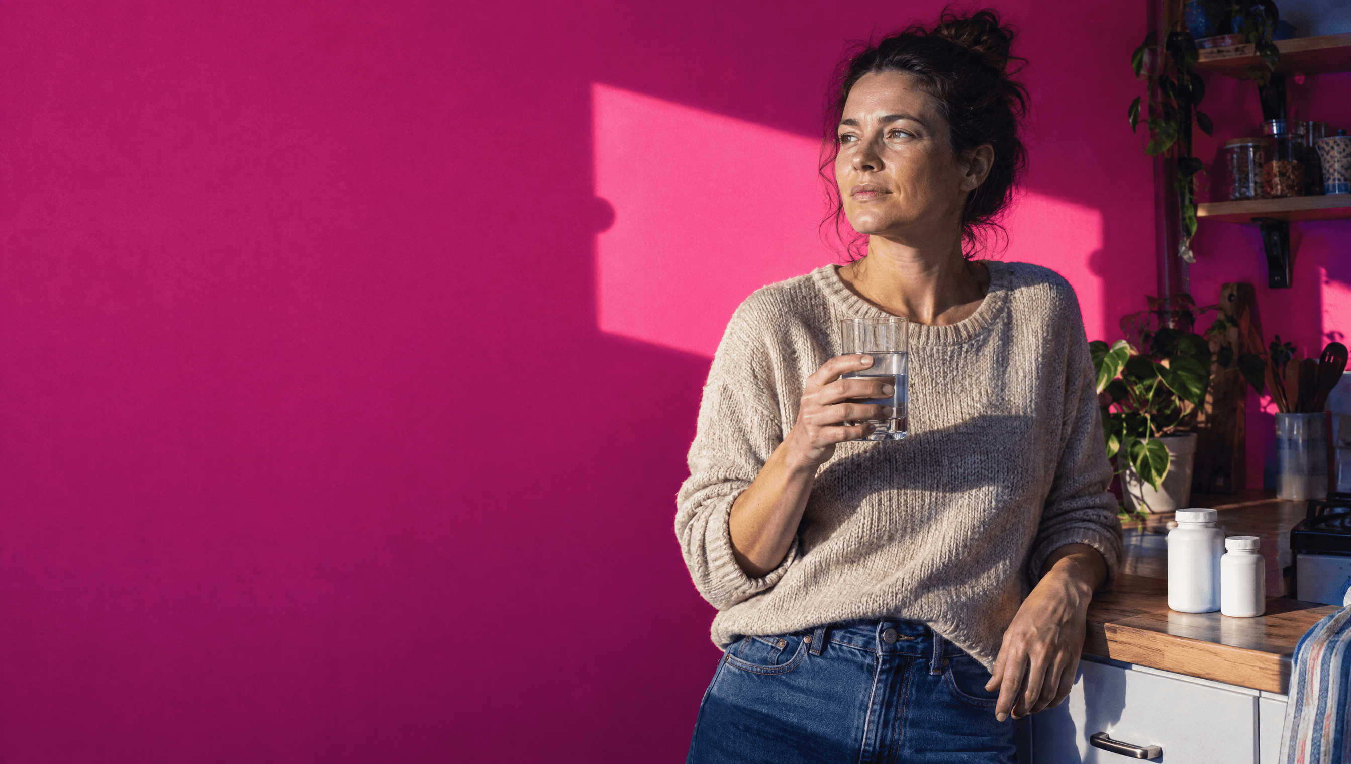 A woman holding a glass of water in a kitchen with a bright pink wall and two plain white bottles on the counter.